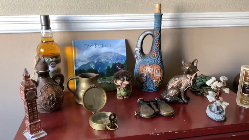 Image of A Diverse Collection of Souvenirs on A Shelf, Including a Scottish Whisky Bottle, a Decorative Jug, Figurines, a Brass Compass, and A Book Titled "The Balkans"