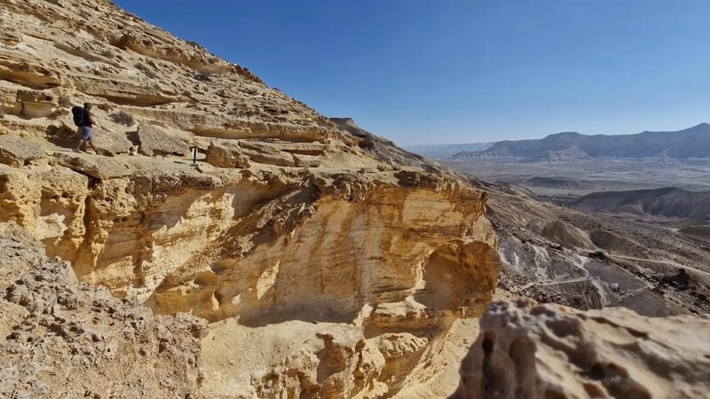 A hiker with a backpack walking along a narrow trail on a rocky cliff in a vast desert landscape
