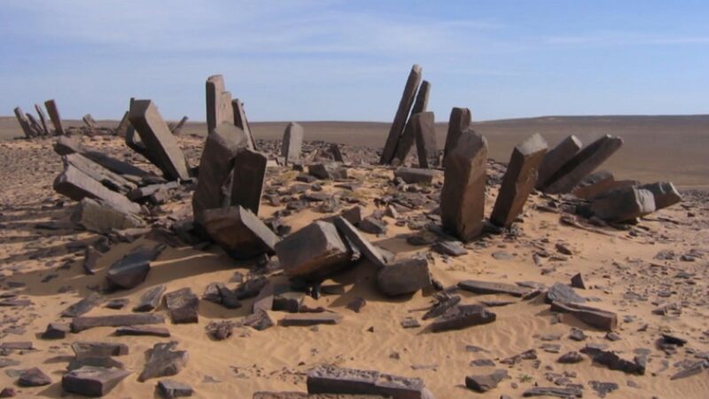 Eroded stone monoliths arranged in a circular formation in the Sahara Desert, partially buried in sand and scattered with debris