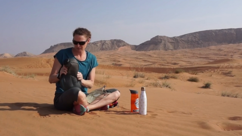 Hiker sits on desert sand with water bottles and gear, resting under the sun in an arid landscape